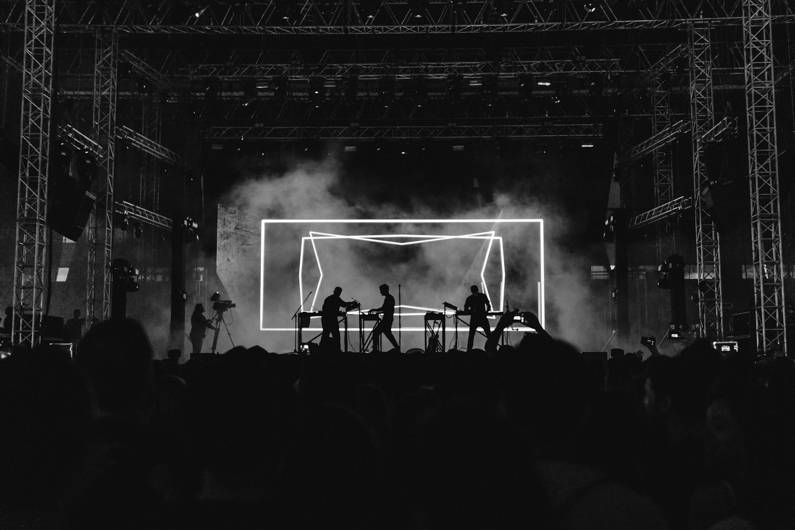 An electronic act silhouetted behind their gear on a smoky stage, performing in front of a glowing geometric light frame backdrop while a crowd watches, the kind of concert visuals a small venue can run from a laptop