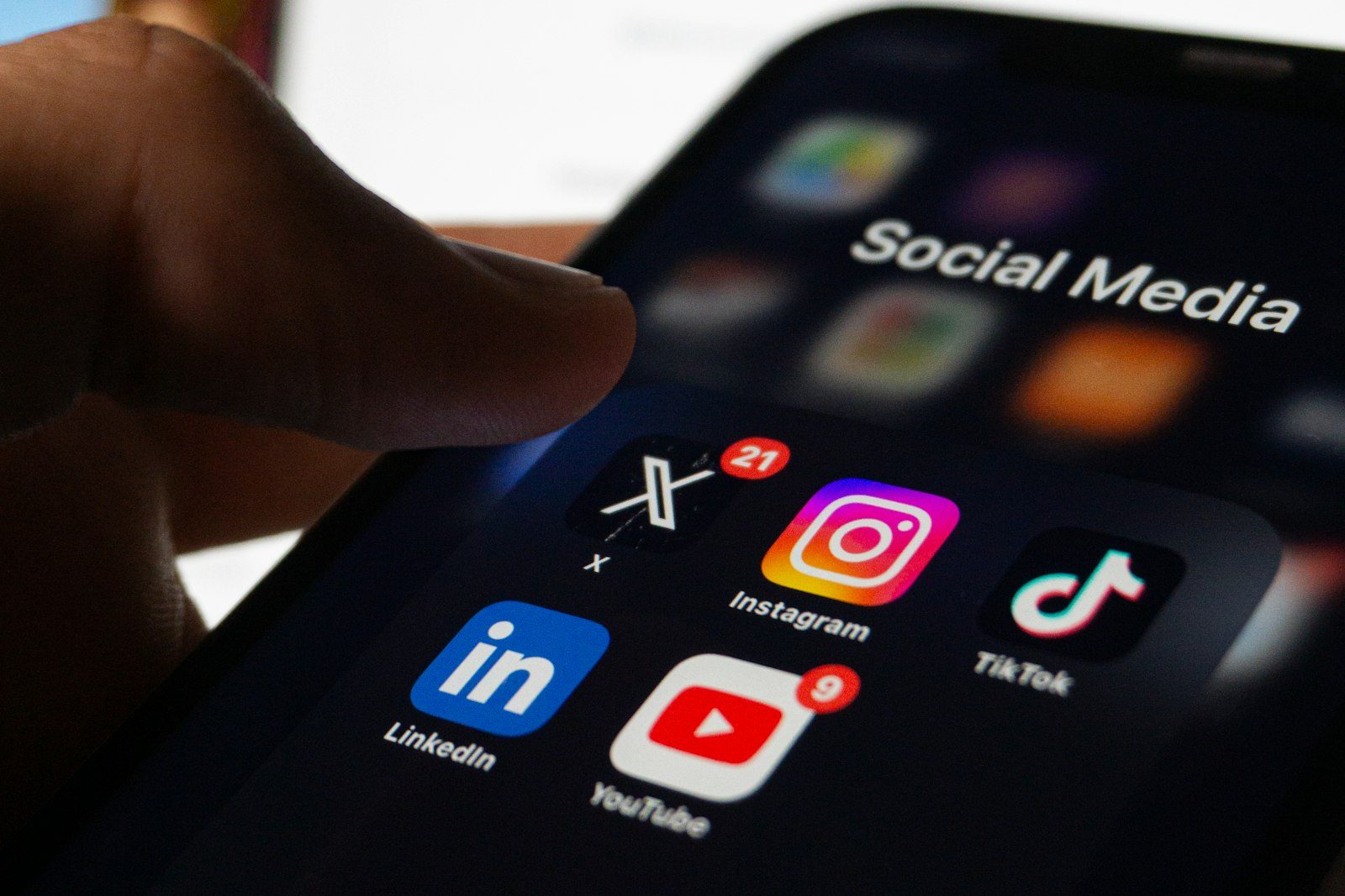 A thumb reaching toward a phone showing a social media app folder with the YouTube, Instagram and TikTok icons