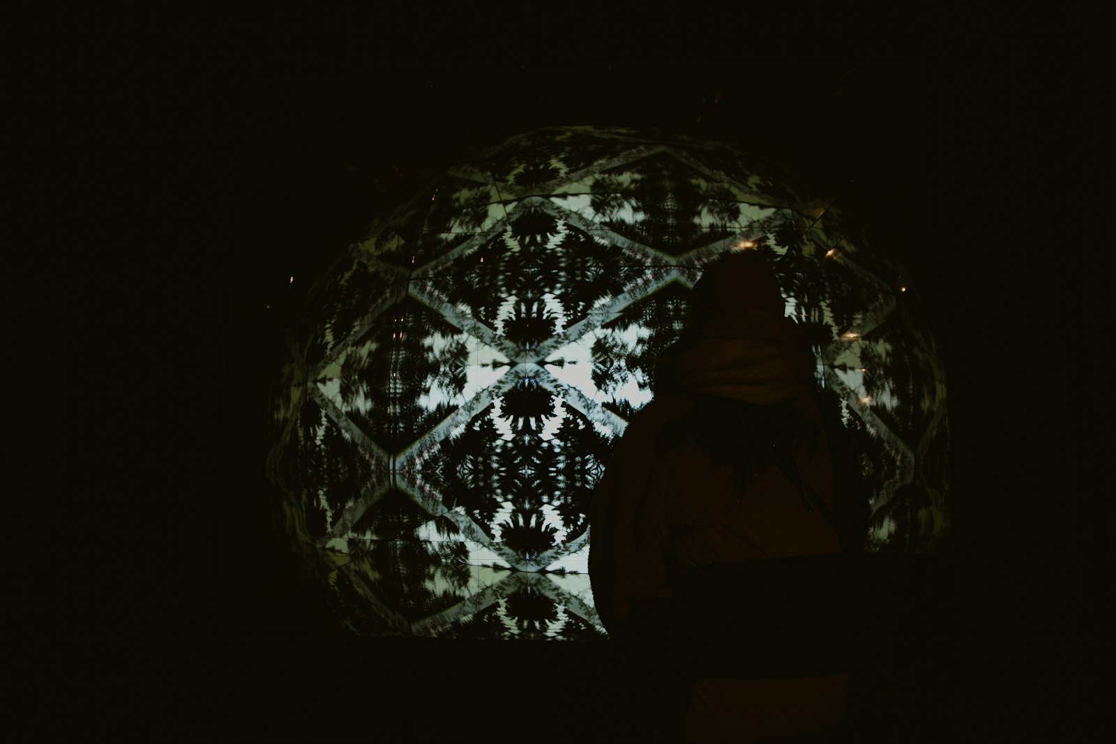 A symmetrical kaleidoscope pattern projected across a dark dome screen while one silhouetted person watches