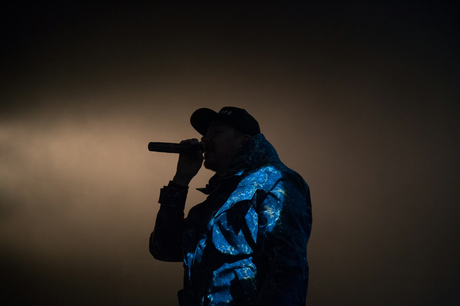 An MC in a snapback holding a microphone, lit blue against haze on a dark stage, the kind of performance a rap visualizer video is standing in for