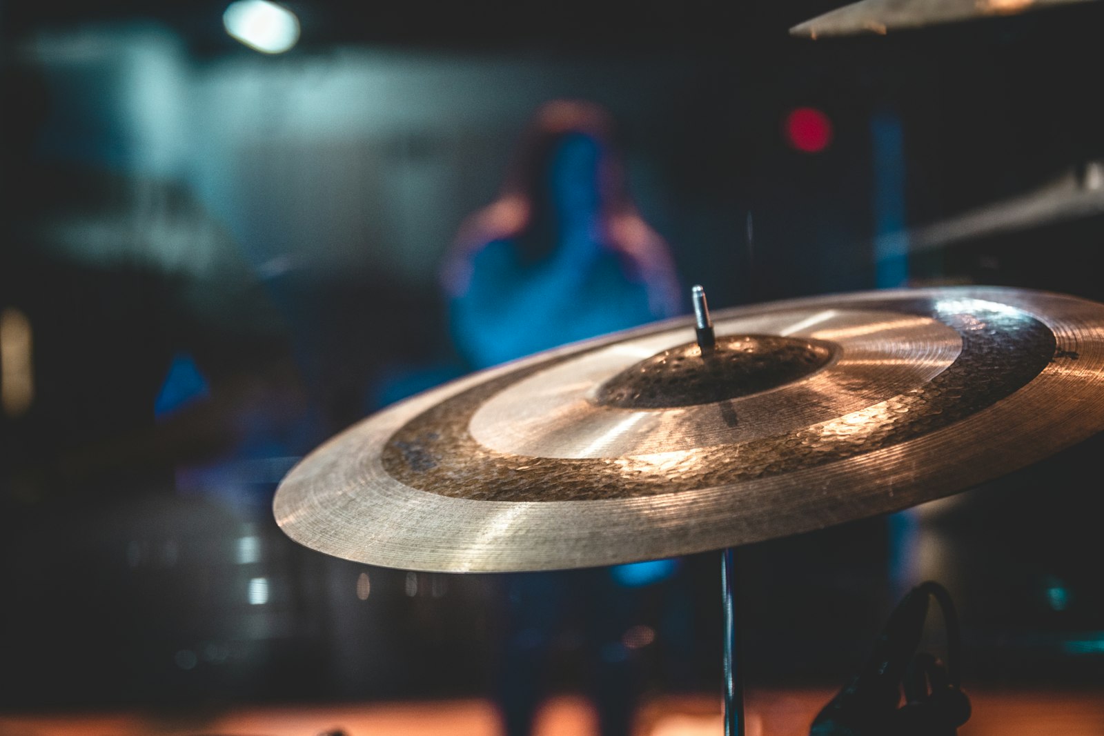 A ride cymbal in close focus on a dimly lit stage, its bell and lathed rings catching warm light, with a musician blurred in blue behind it