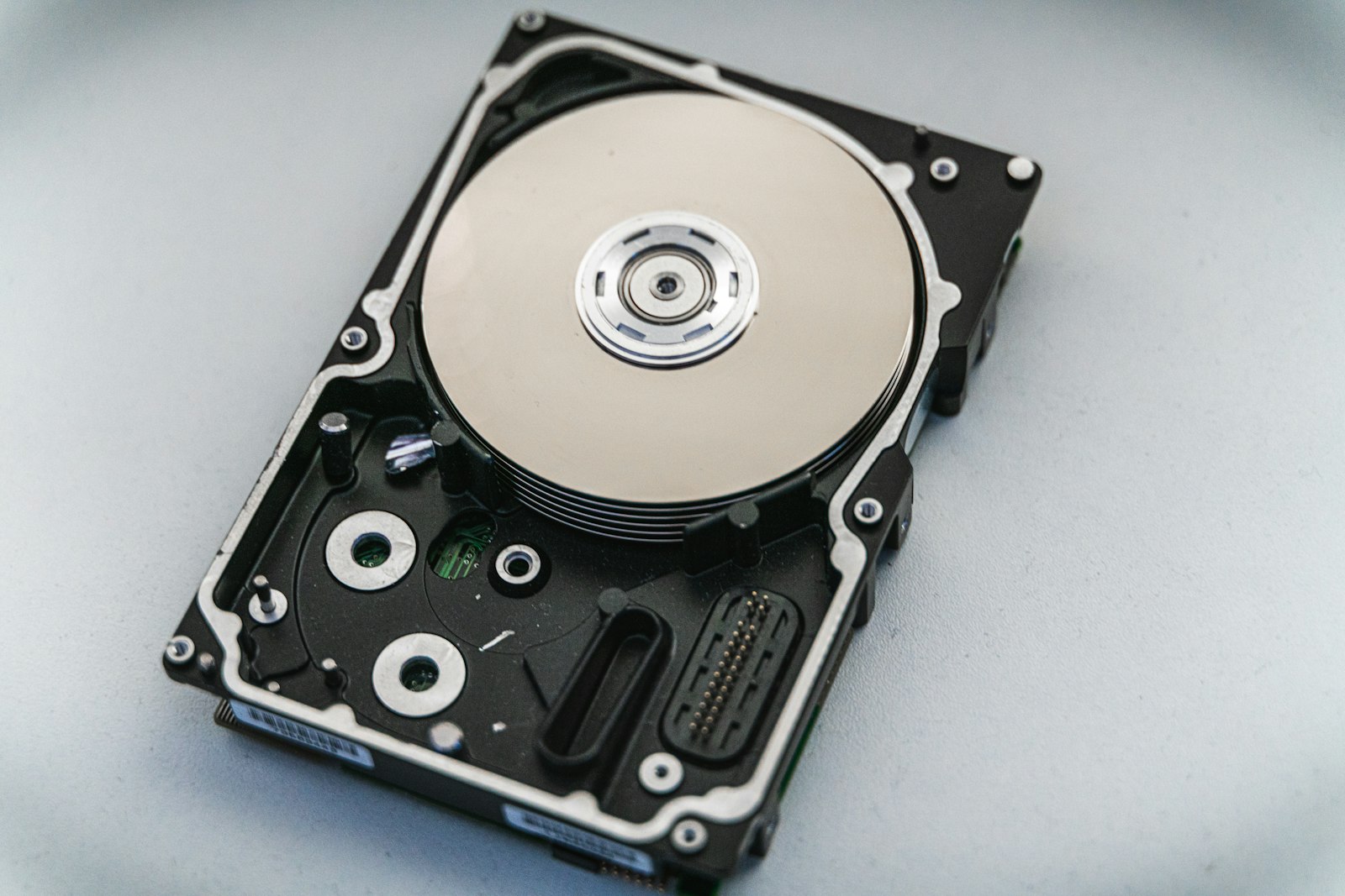 An opened 2.5 inch hard disk drive lying on a pale surface with its casing removed, the mirrored platter and read head exposed, local storage of the kind an unreleased track sits on