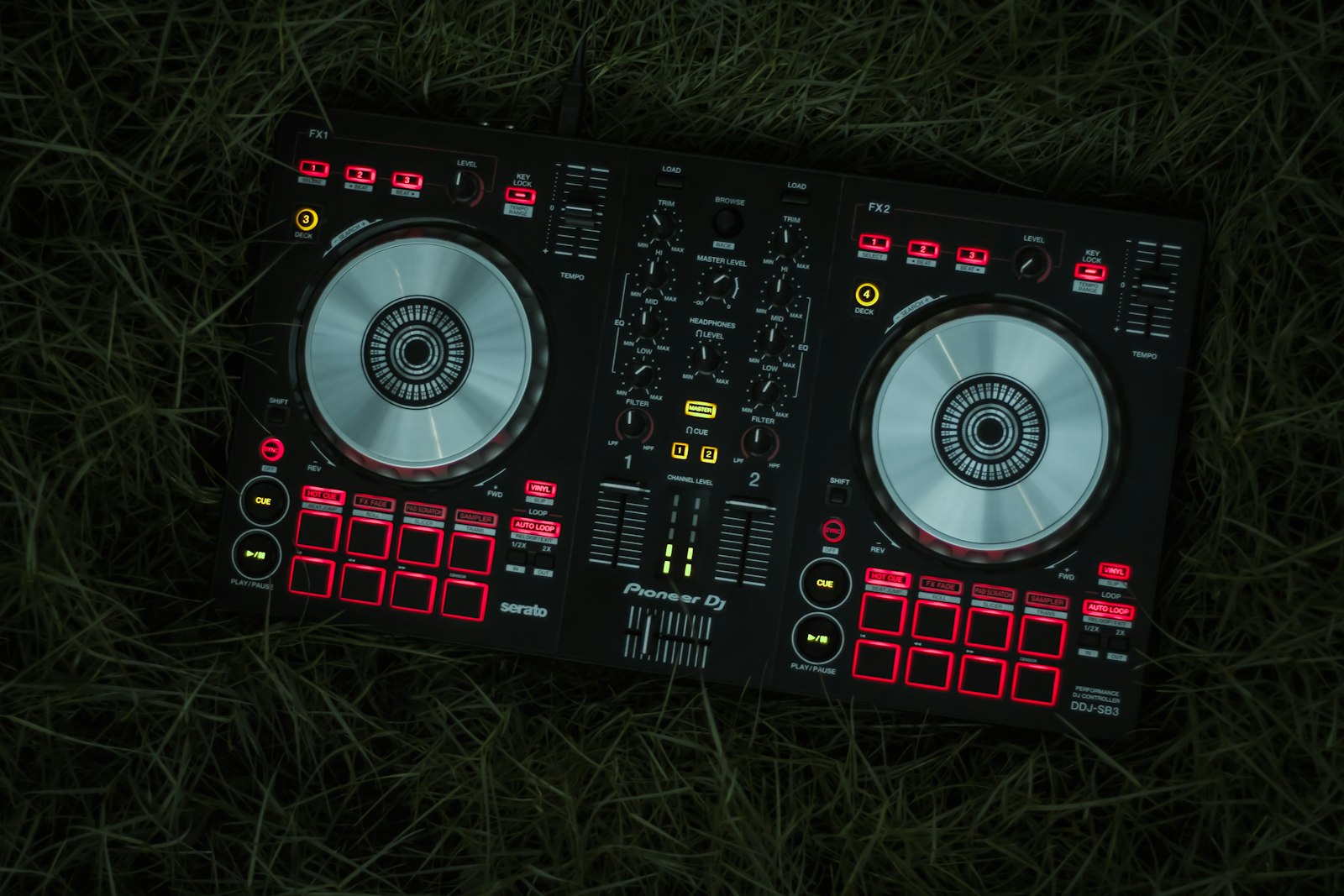 A Pioneer DJ controller lying in grass lit deep red, its two TEMPO faders and SYNC buttons picked out along the top edge